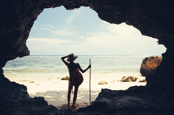 Silhouette of young woman standing in cave on the beach with hat, stick and backpack
